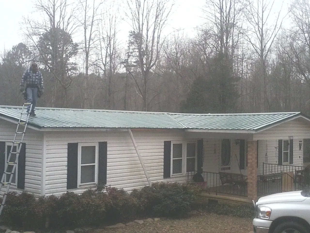 Worker on a ladder beside a new green metal roof on a ranch home in Mill Creek