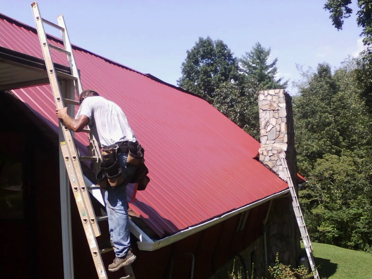 Roofer on a ladder beside a red metal roof and stone chimney — Soffit & Fascia Repair by Roofing Brothers in Mill Creek